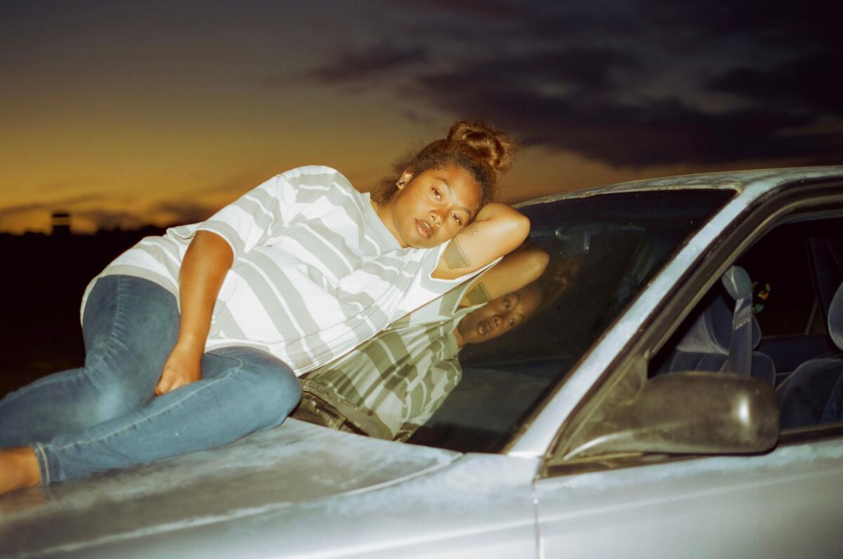 A preoccupied woman sits on the hood of a car, leaning against the windshield.