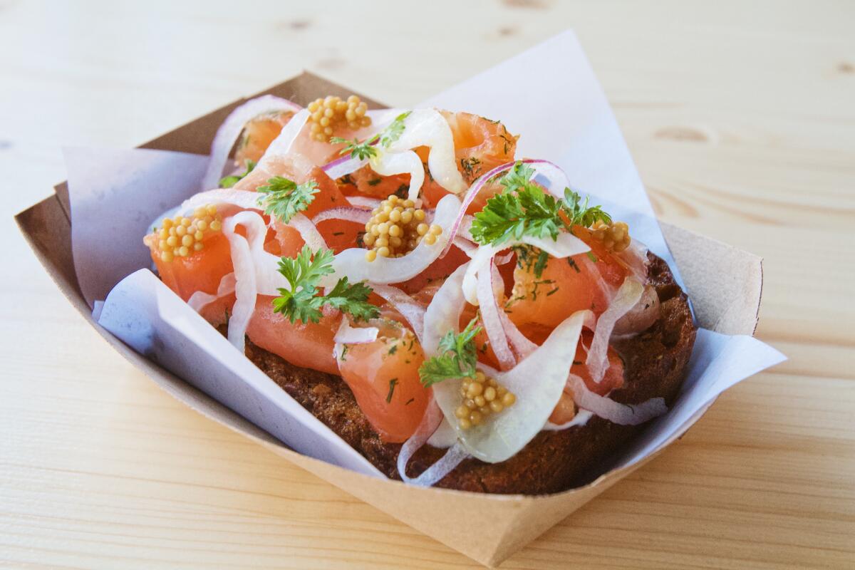 Close-up of a cured trout tartine, decorated with mustard seeds and shaved fennel, on a cardboard plate.