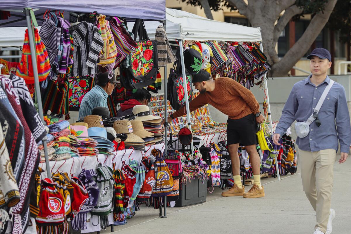 One man walks on a sidewalk alongside a street vendor stall, while two vendors sell scarves and other items.