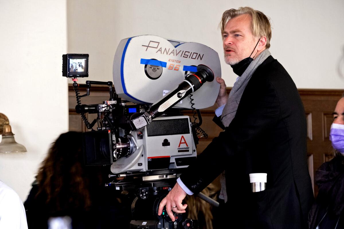 A blond man, a face mask under his chin, stands next to a Panavision movie camera