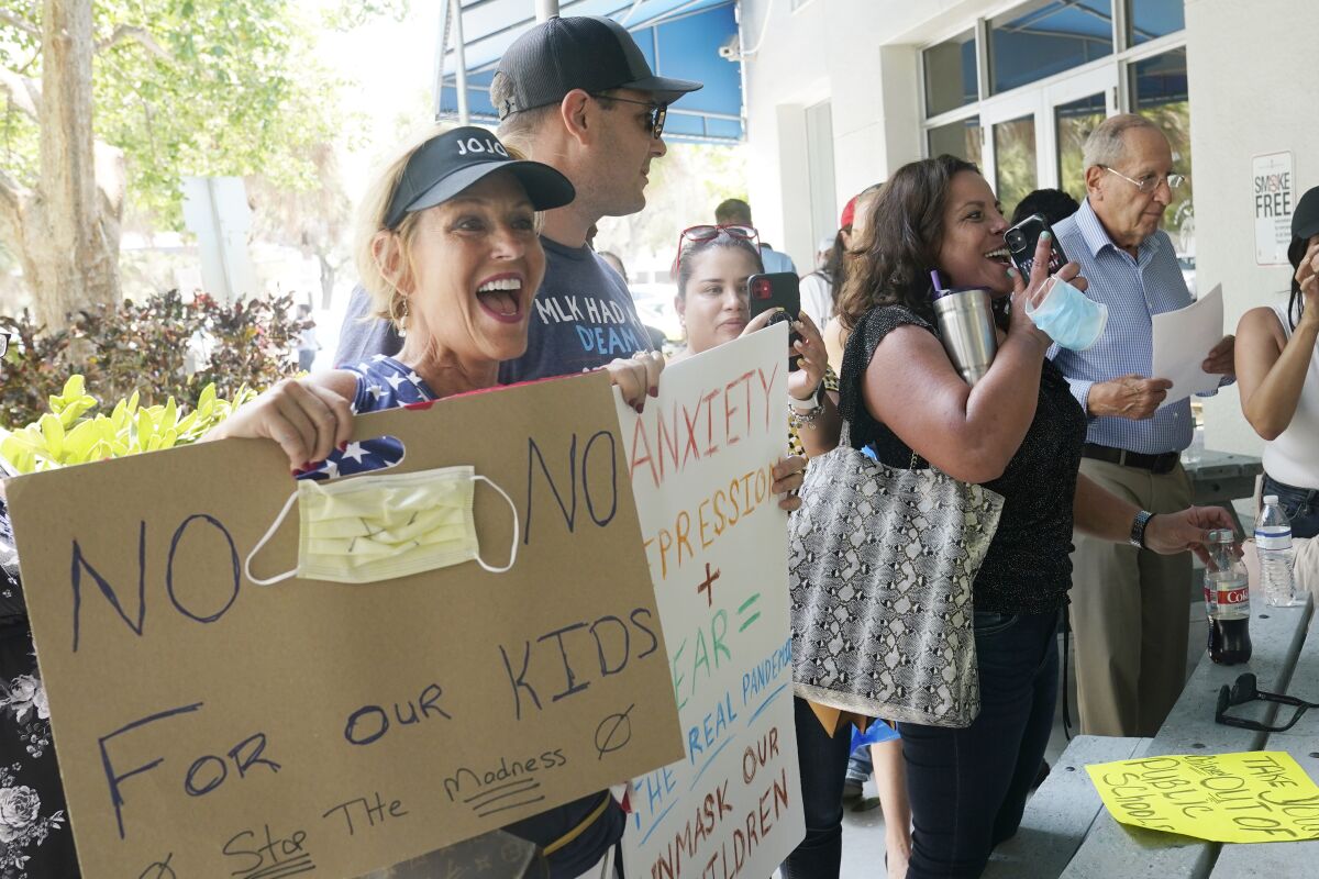 A woman stands with others outside a building, holding a sign that says "No masks for our kids. Stop the madness."