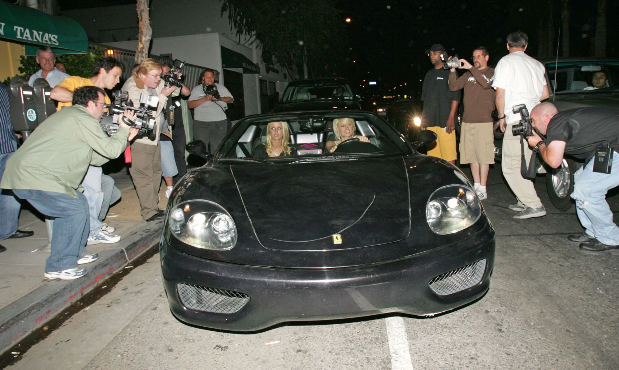 Nicky and Paris Hilton driving a black sports car with lines of photographers on either side of them