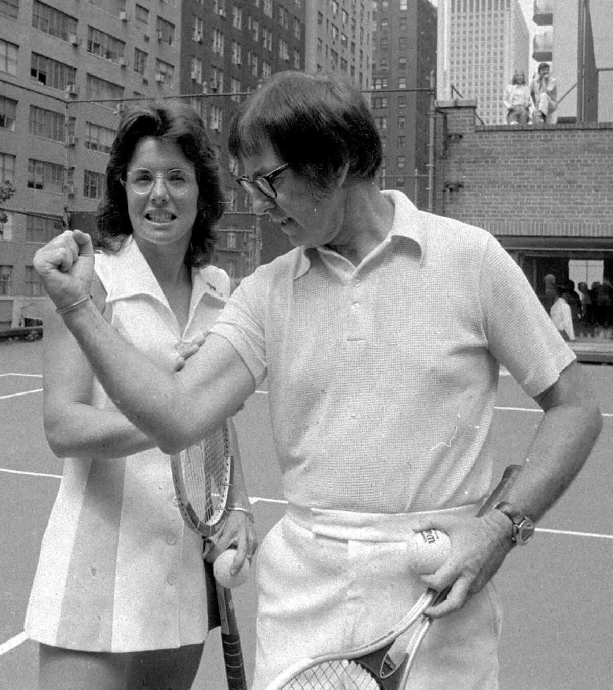 Commentary: She left faculty to overcome tennis. At 81, Billie Jean King is again, chasing a level 2 A woman with dark hair and glasses, holding a racket, next to a man flexing his arm, with a racket and ball in his other hand