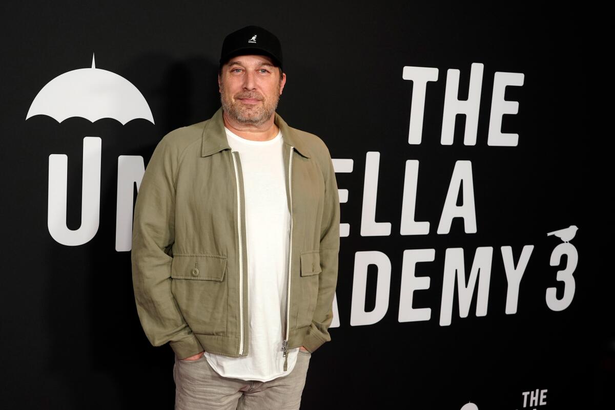A man in a tan jacket, white T-shirt and black hat standing with hands in pockets in front of an 'Umbrella Academy' backdrop