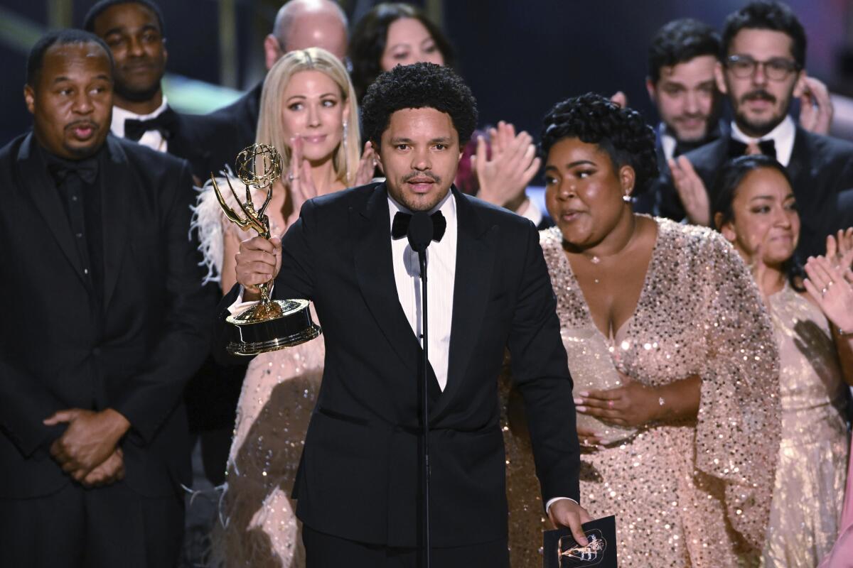 Trevor Noah holds an Emmy award in the air and speaks into a microphone with cast and crew surrounding him. 