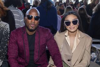 A bald Black man in a maroon suit and sunglasses sitting with an Asian woman with long hair in a beige suit and sunglasses