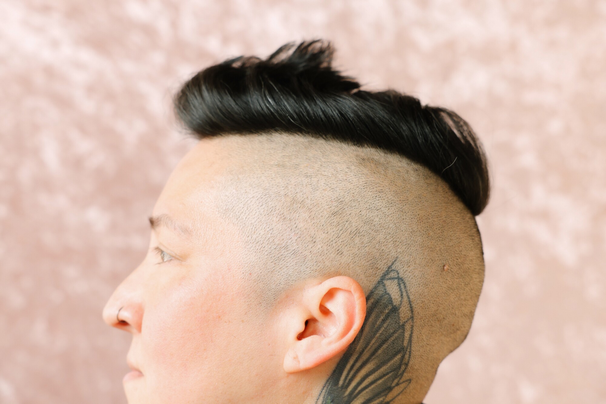 A person has shaved hair on the side of his head, has long hair on top and a tattoo near the ear