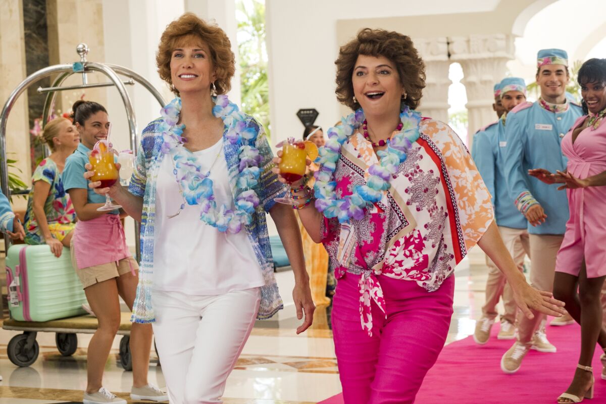 Kristen Wiig and Annie Mumolo walk through a hotel lobby wearing leis and holding tropical cocktails.