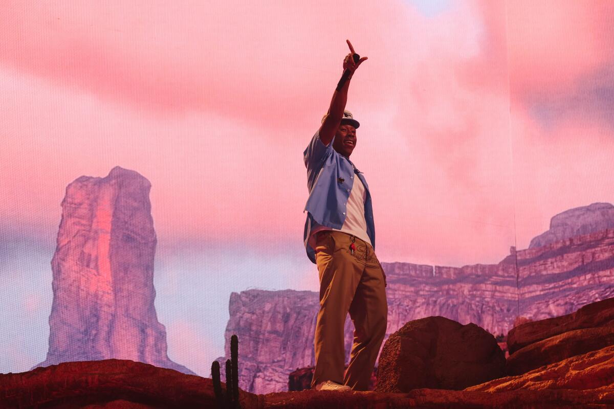 Tyler, the Creator, holding up one hand and pointing to the sky, on a stage made to look like a rocky desert landscape