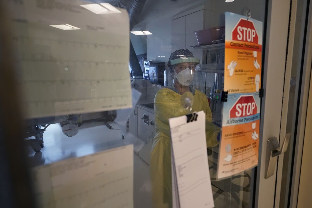 Behind a closed glass door, a hospital nurse wears a protective yellow gown and face shield