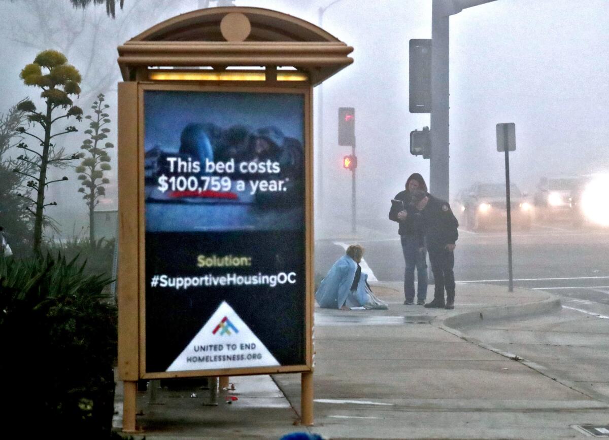 Mike Brumbaugh, right, Costa Mesa senior code enforcement officer, and Costa Mesa neighborhood improvement manager Stacy Lumley speak with a homeless man who was sleeping on the sidewalk at Victoria Street and Harbor Boulevard on Feb. 26. The sign on the bus shelter promotes supportive housing, which combines housing with services and connections to needed resources.