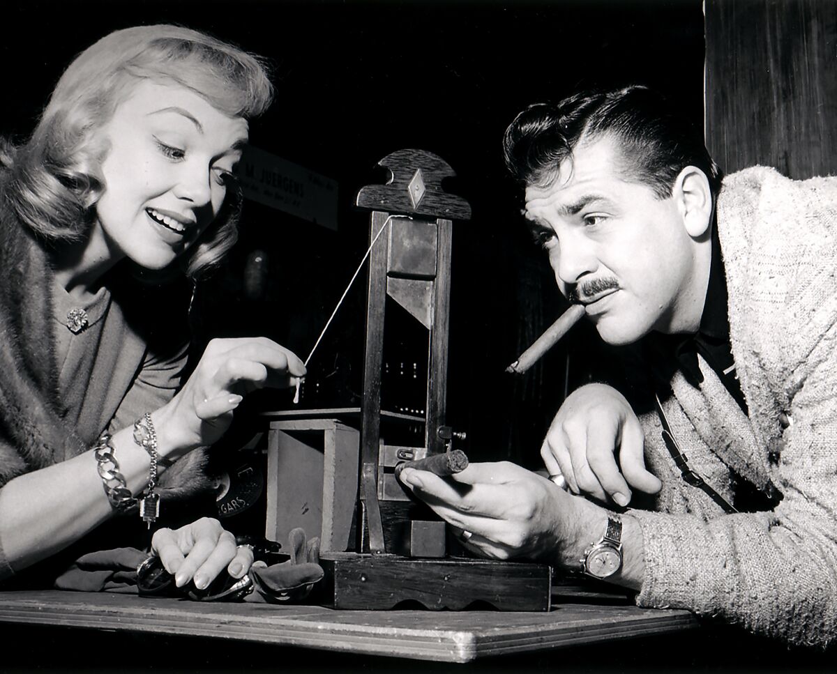 Edie Adams holds the string of a tiny guillotine as Ernie Kovacs inserts a cigar to cut.