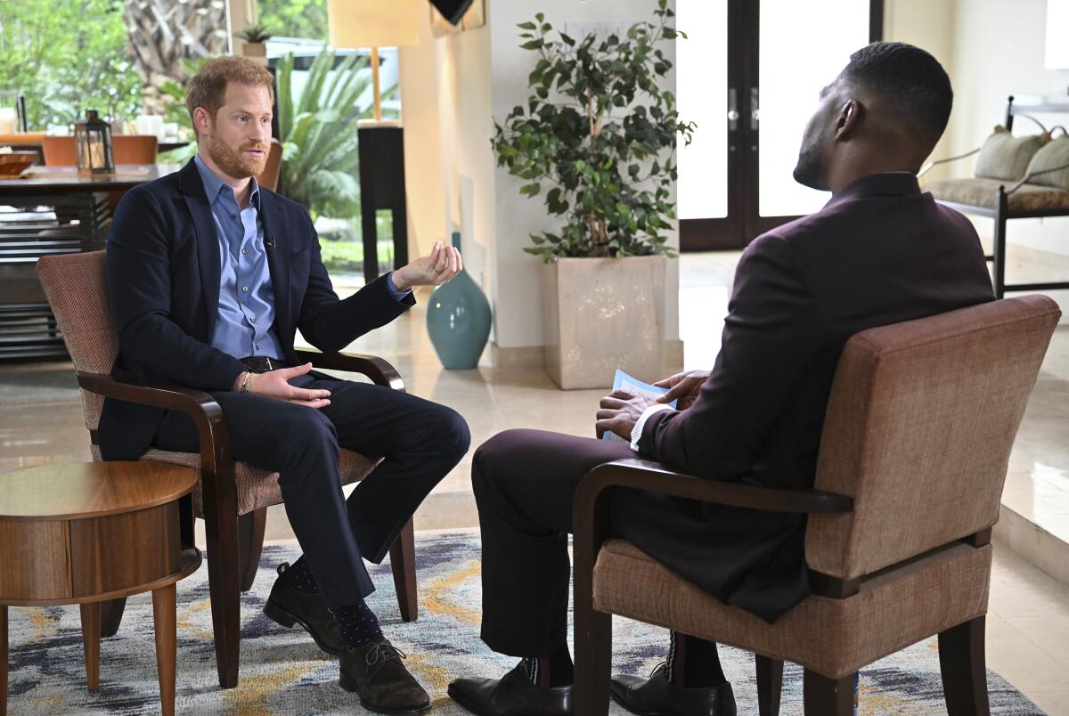A man with red hair and a beard sits and talks with a black-haired man whose back is to the camera.