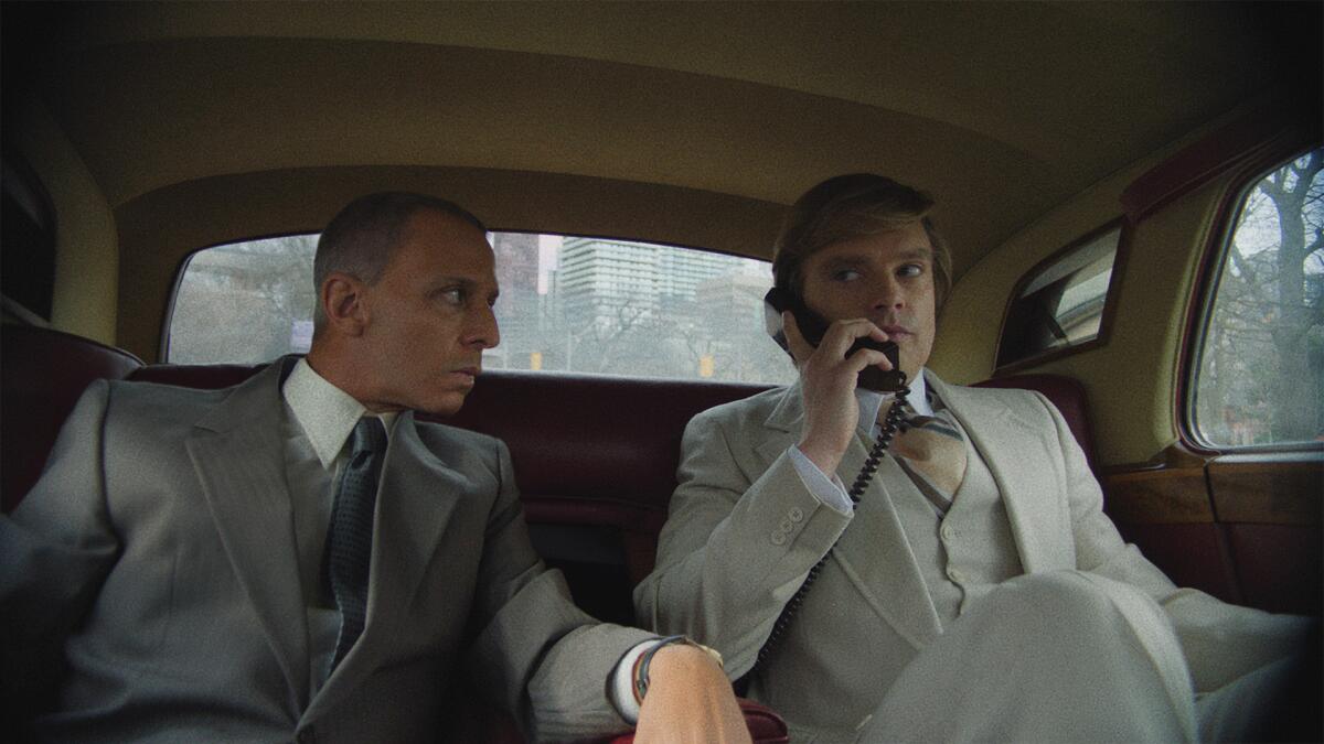 Two men in suits in the back seat of a limo, one holding a corded phone handset.