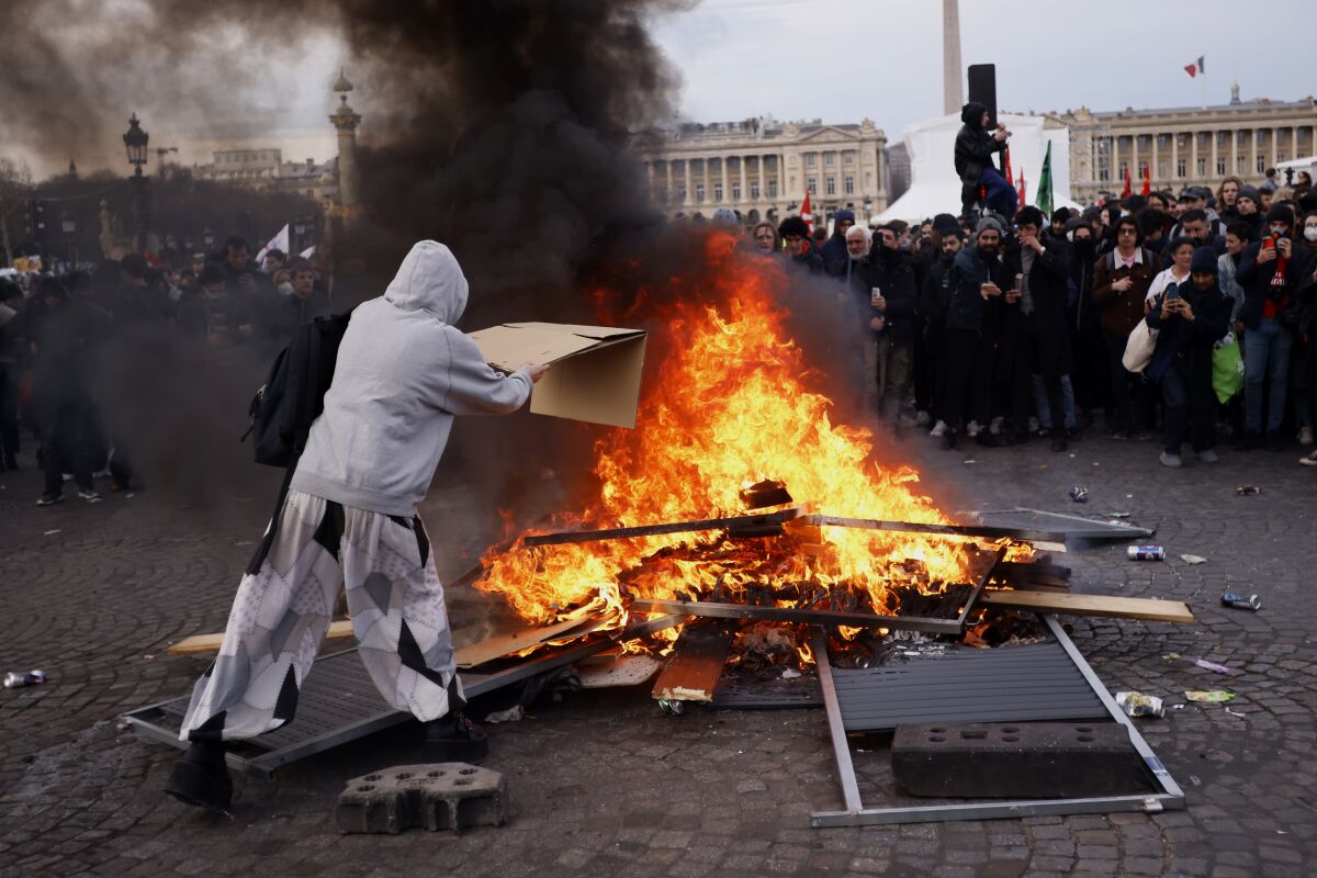 A protester throwing cardboard into a pile of burning pallets in a city square as a crowd looks on