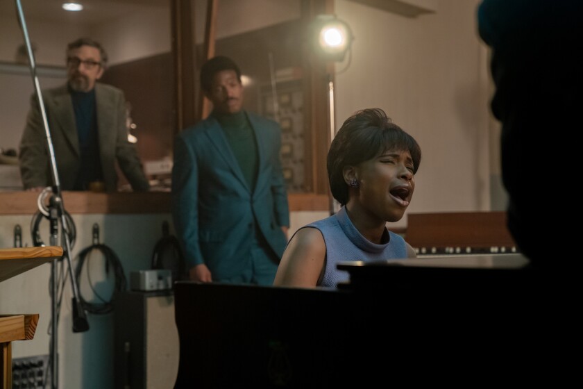 Two men stand behind a woman seated at a piano and singing.