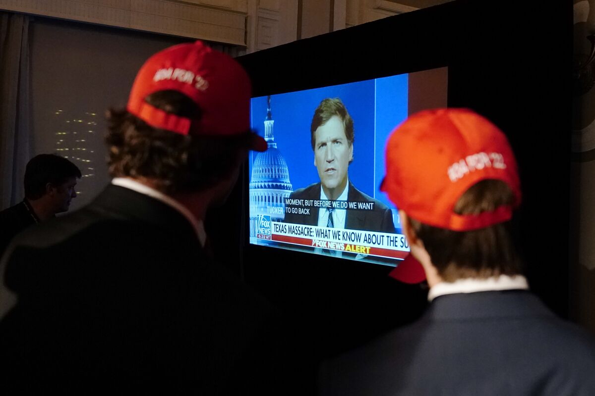 Two men in red baseball caps watch Tucker Carlson on a television screen.