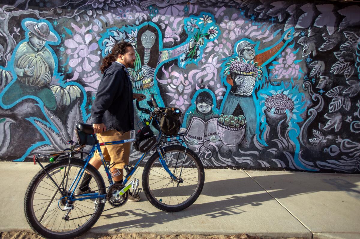 A Latino man with a ponytail walks a bicycle in front of an ornate mural picturing street vendors and a farmworker