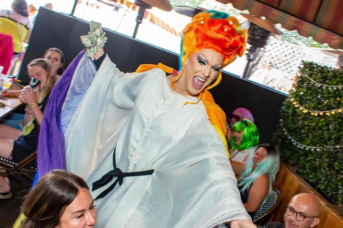 An orange-haired drag queen stands behind a seated restaurant diner