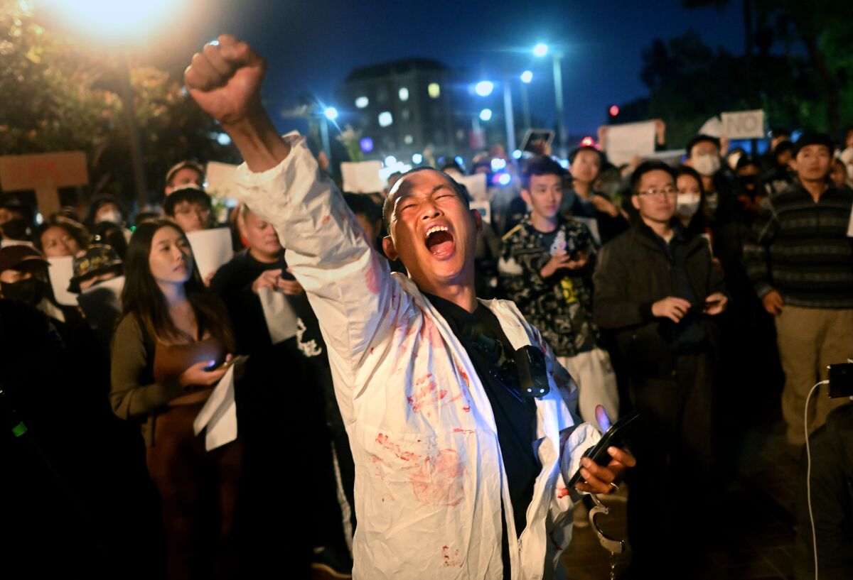 A person with a buzz cut and a white jumpsuit with bloody handprints raises a fist in   protest