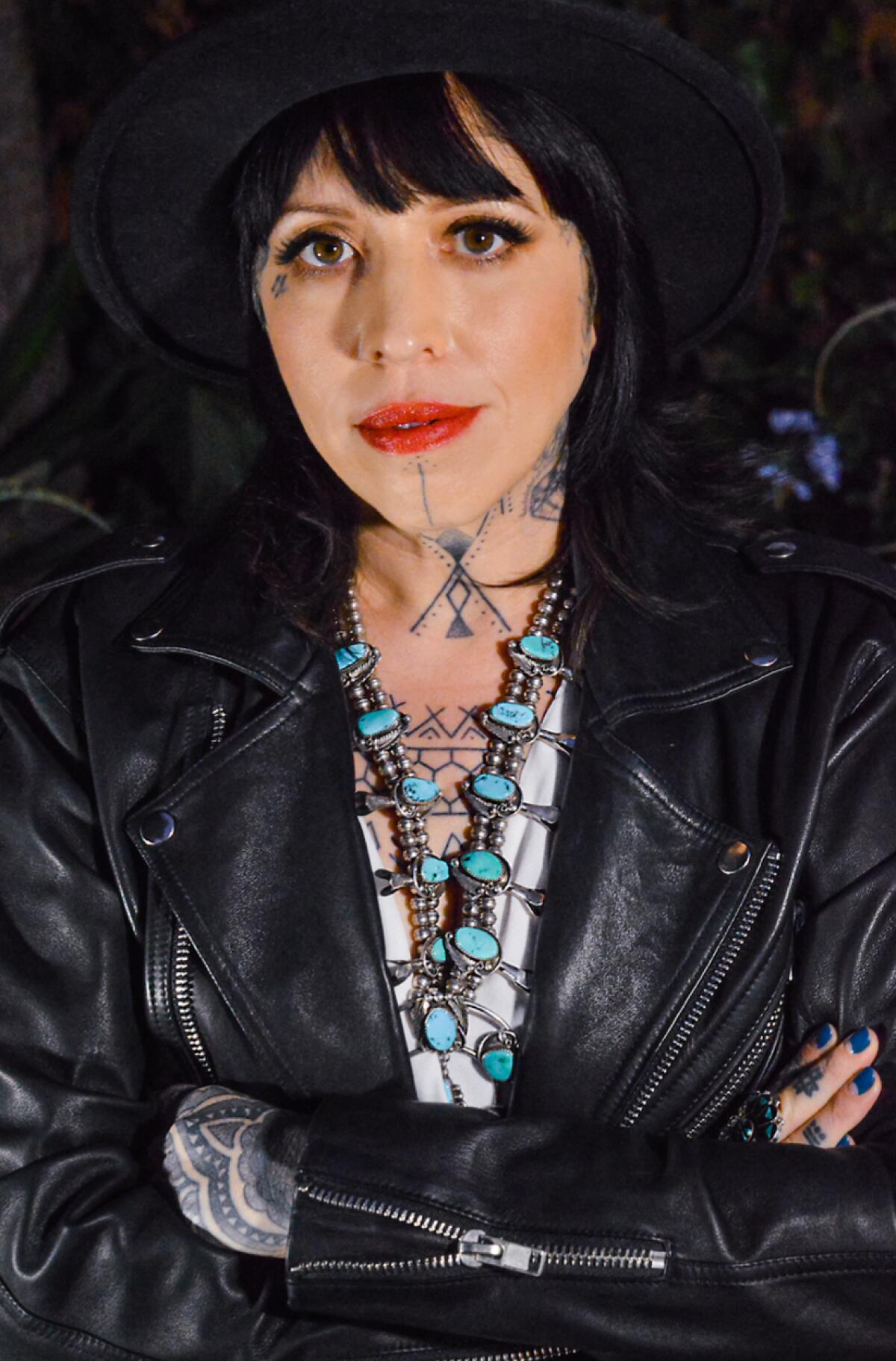A woman with neck tattoos and turquoise necklace wears a black leather jacket.