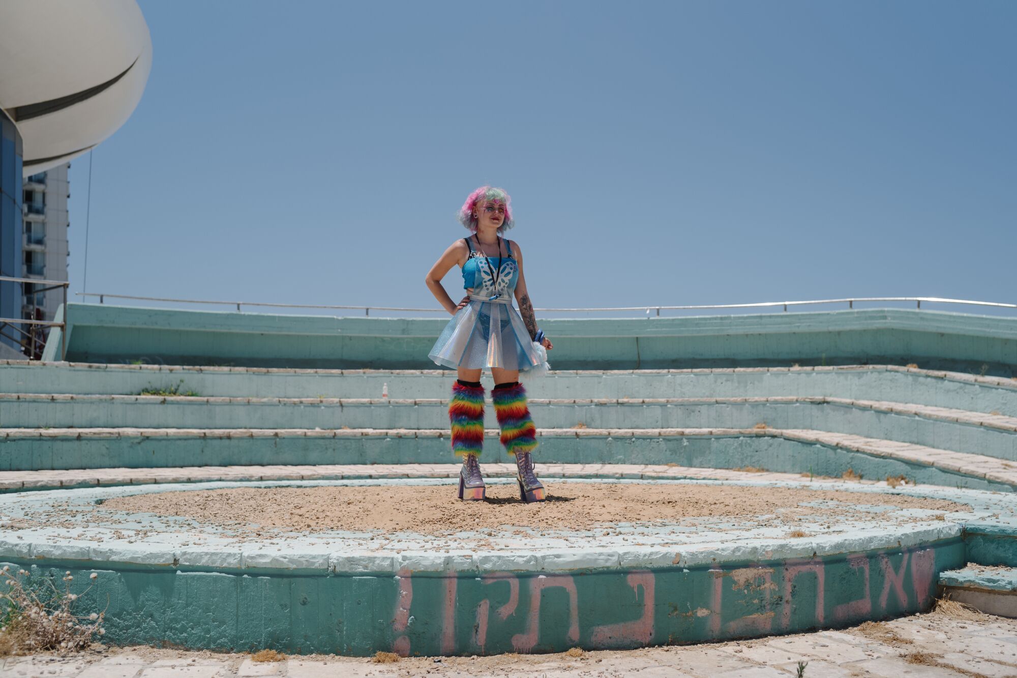 A woman with platform shoes and rainbow hair and pants stands near the beach