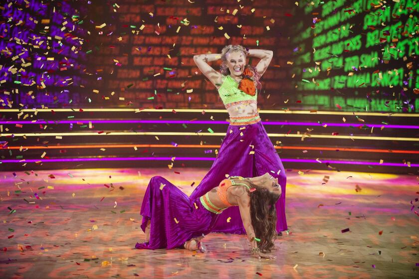 A blond woman in vibrant outfit poses behind a brunette woman in a matching outfit, surrounded by confetti