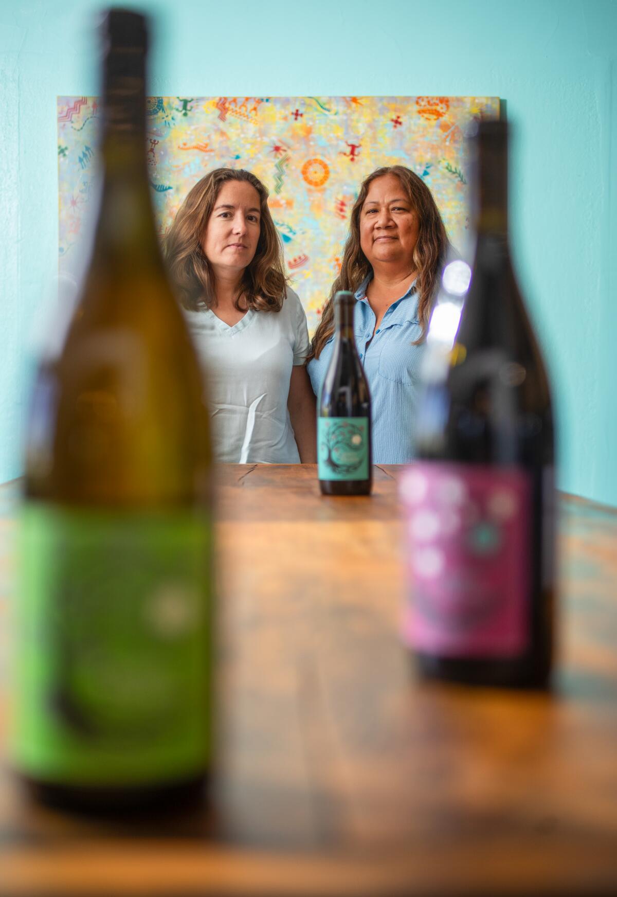 Two women at the far end of a table with wine bottles on it