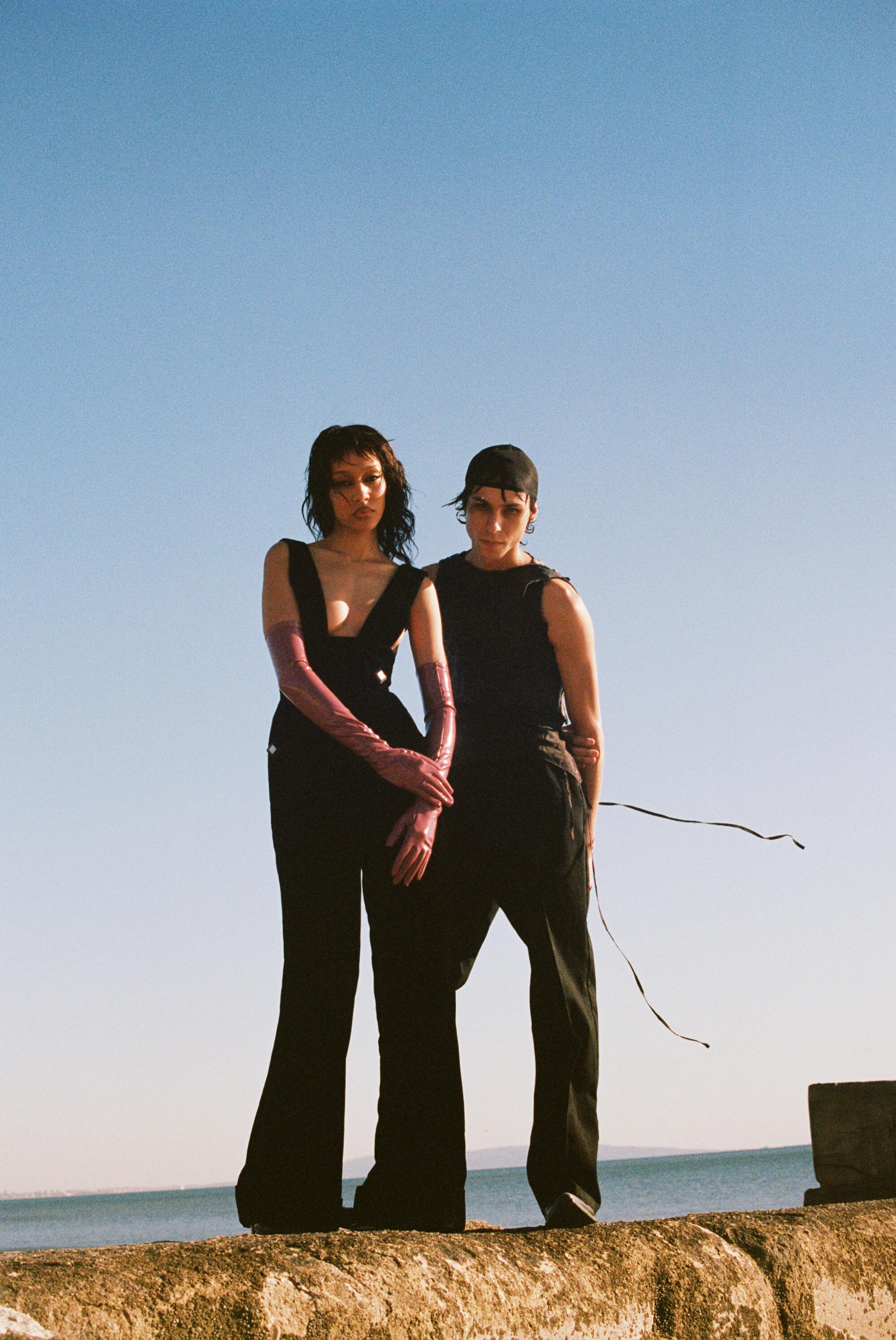 Two models in black outfits stand on a rock wall.