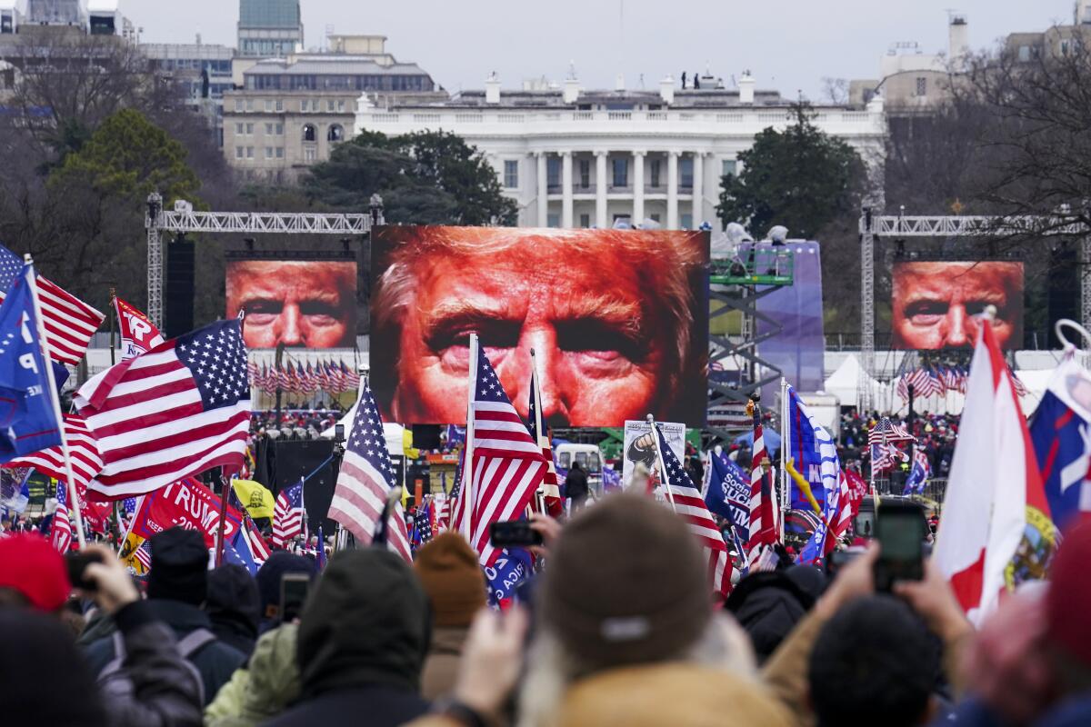 A crowd with American flags and Trump signs views an extreme closeup of his face on large screens outside the White House