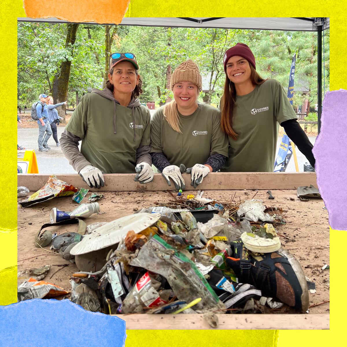 Three women in matching T-shirts, beanies and work gloves smile for a photo behind a pile of trash.