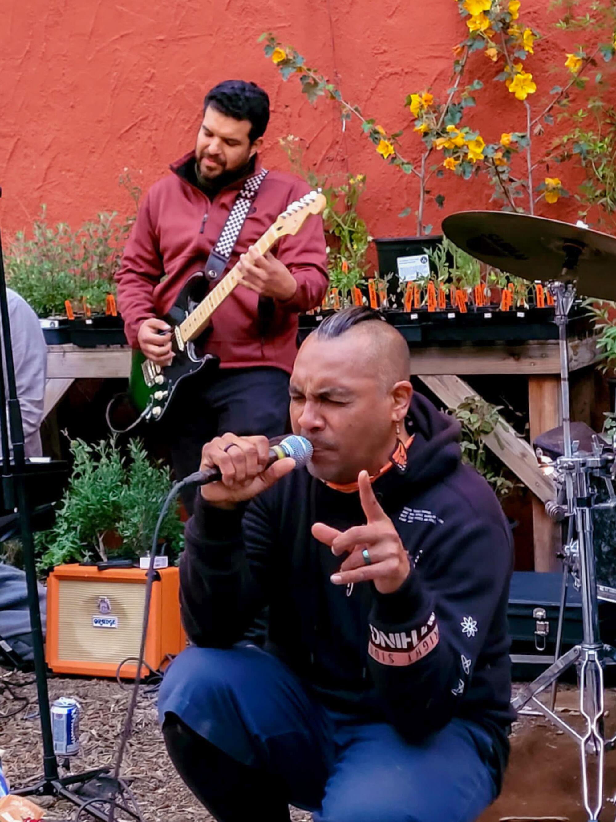 Frontman Antonio Sanchez crouches while singing into a microphone with lead guitarist Rico Ramirez playing behind him.