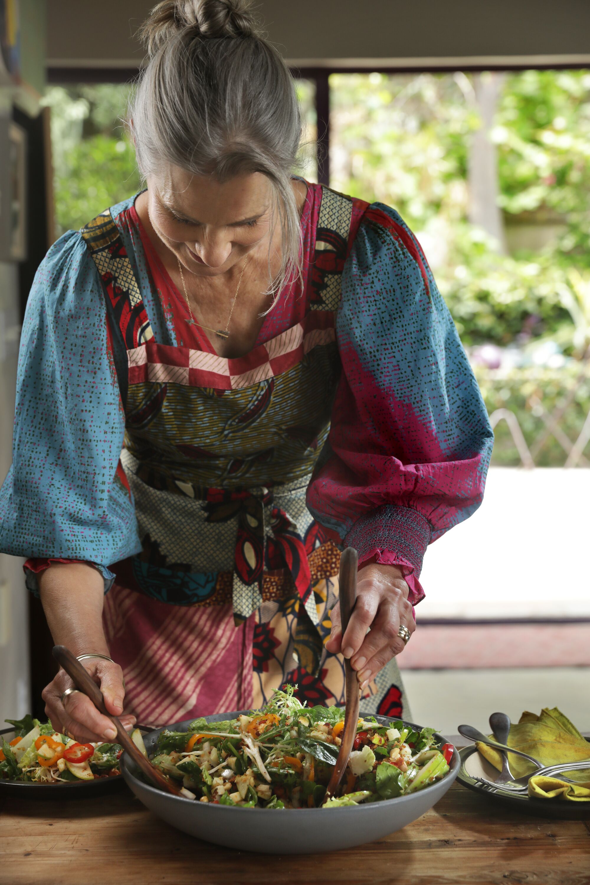 A woman in a kitchen leans over a bowl of salad on a counter.