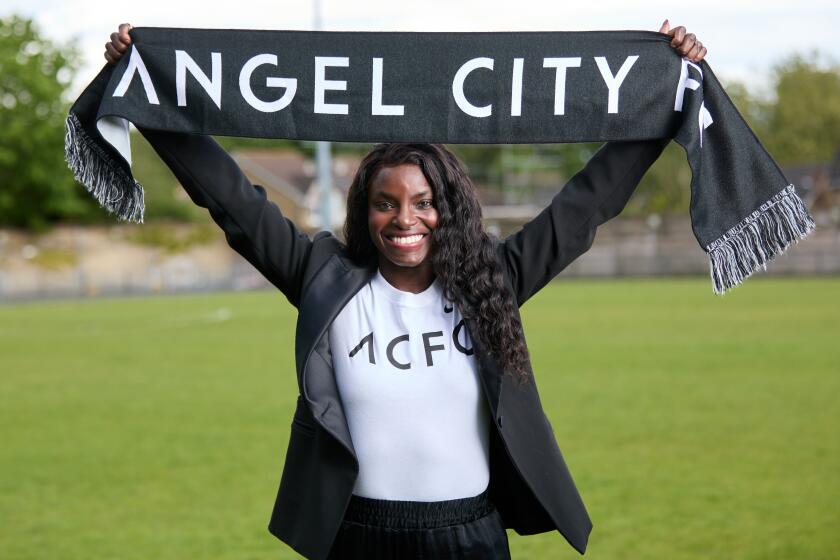 Eniola Aluko holds an Angel City scarf over her head while standing on a soccer field 