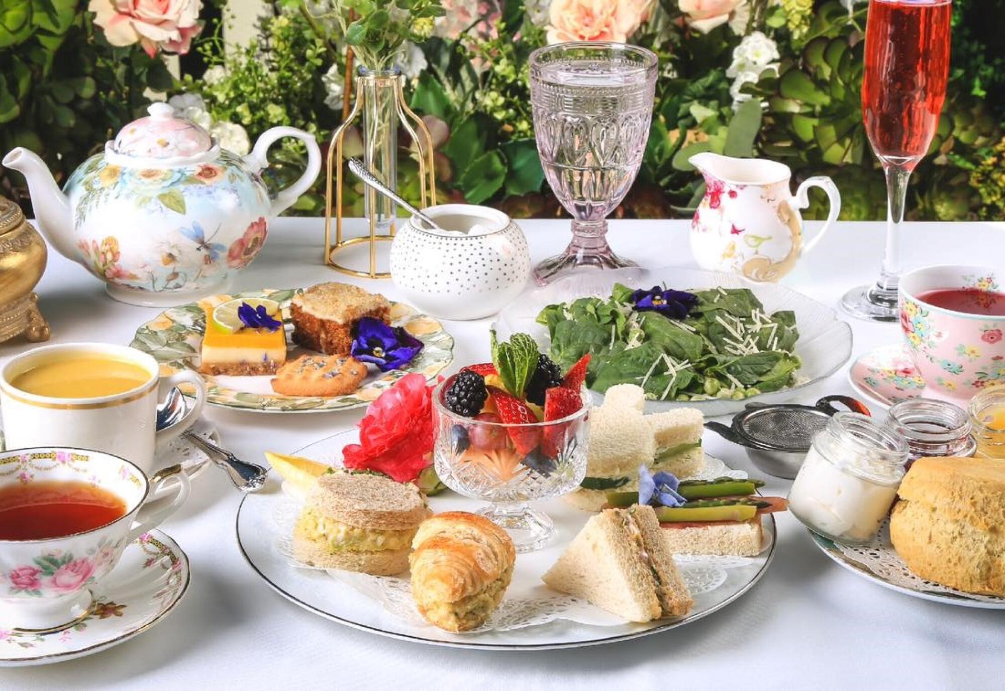 A spread with tea sandwiches, pastries, salad, teacups, a teapot, a glass of water and champagne.