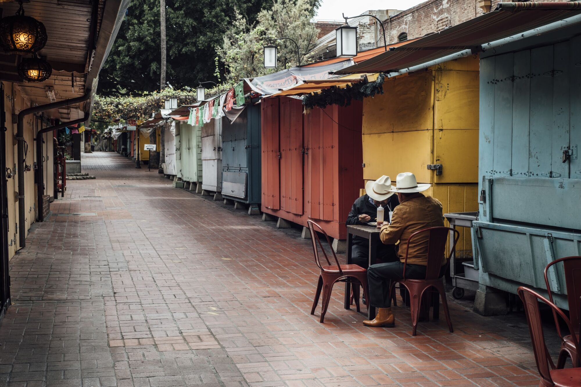 Two men in cowboy hats sit at a solitary dining table outside closed stalls on Olvera Street