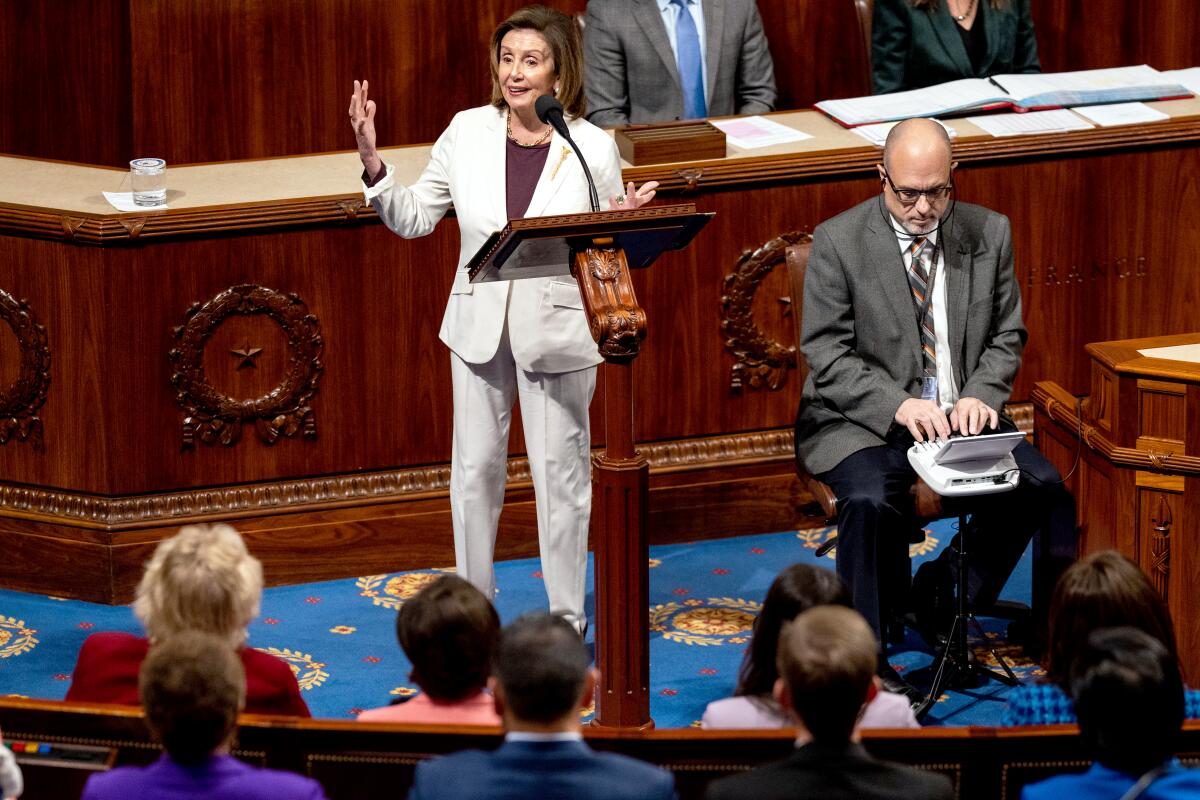 A woman in a white pantsuit stands at a lectern speaking into a microphone. People are seated in rows in front of her.