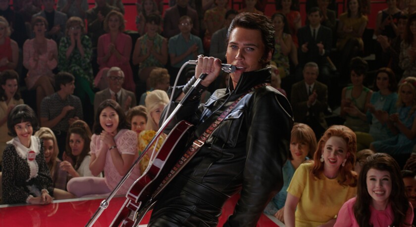 A man in a leather jacket with a guitar leans back as he sings into a microphone in the movie "Elvis."
