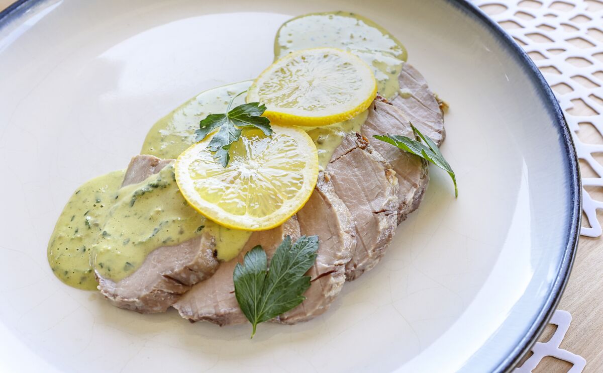 A tuna sauce and lemon slices top pieces of pork tenderloin on a plate.