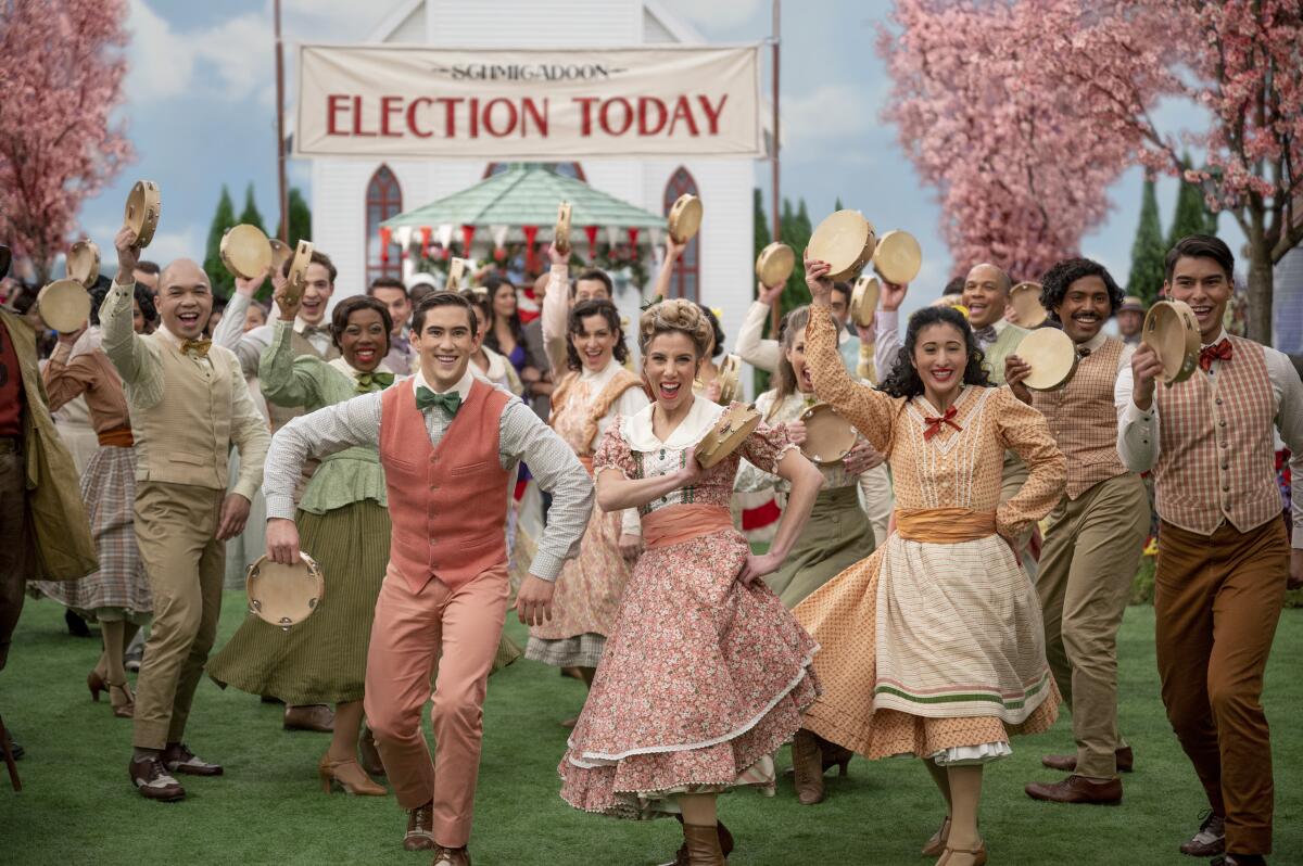 A group of citizens in an old-timey town sing and dance before an Election Day banner.