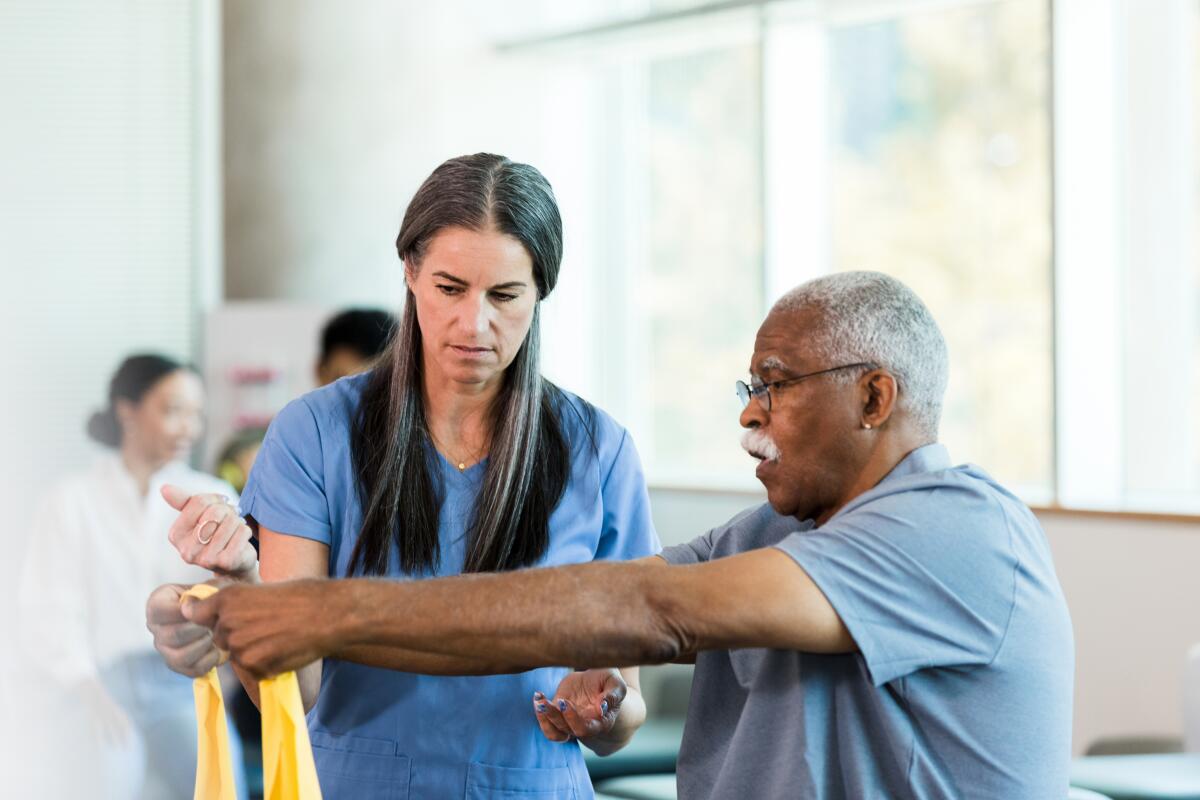 A physical therapist guides a senior adult man as he grips and stretches an elastic band.
