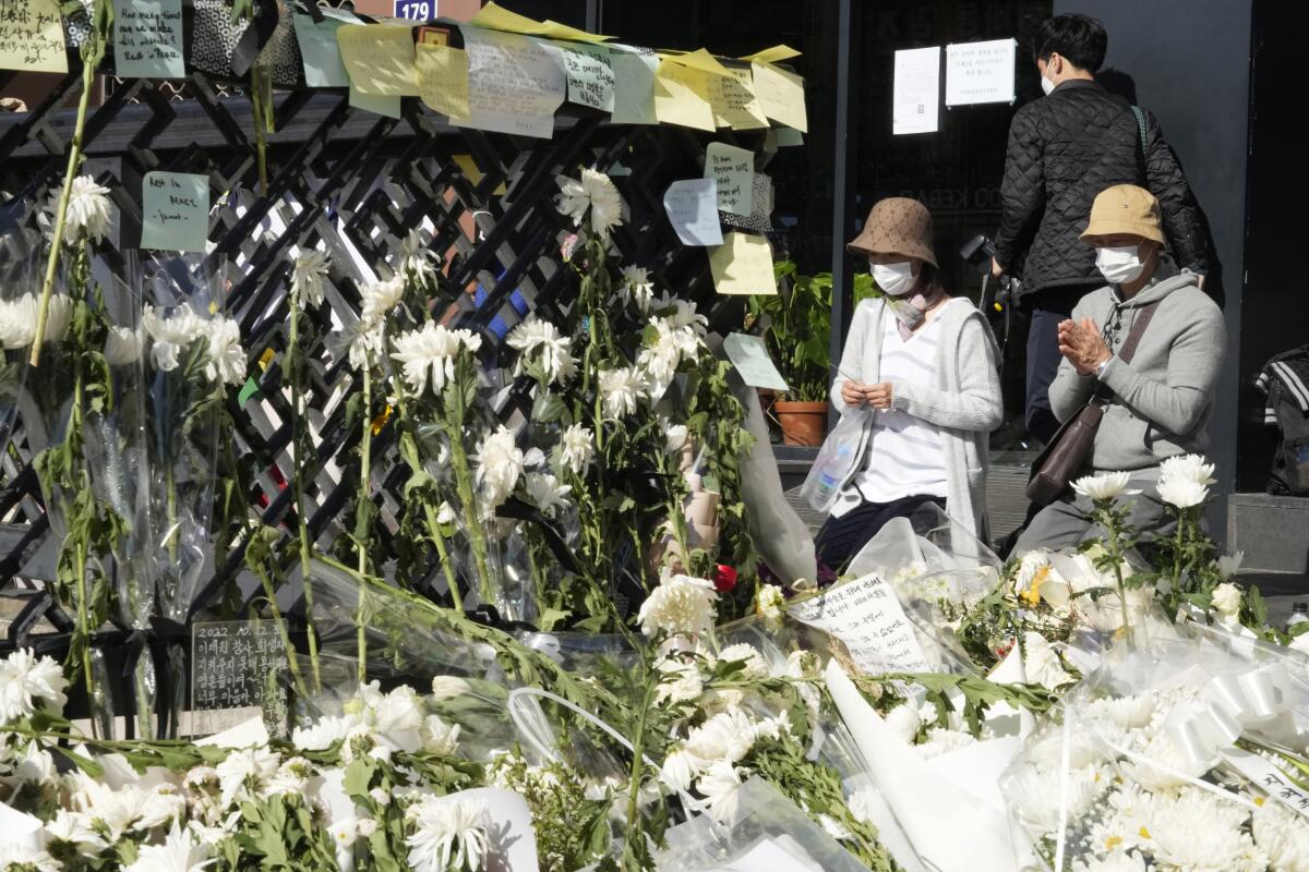 Two people wearing bucket hats stand, with their hands together, before piles of white flowers 