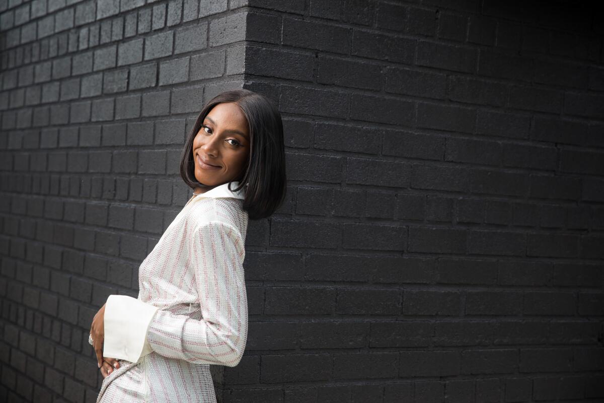 Ziwe poses in a white striped dress with large cuffs against a black brick wall.