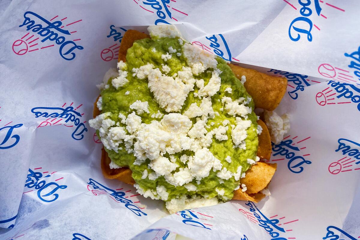 An overhead photo of taquitos under avocado salsa and cheese, sitting in Dodgers-branded wax paper.