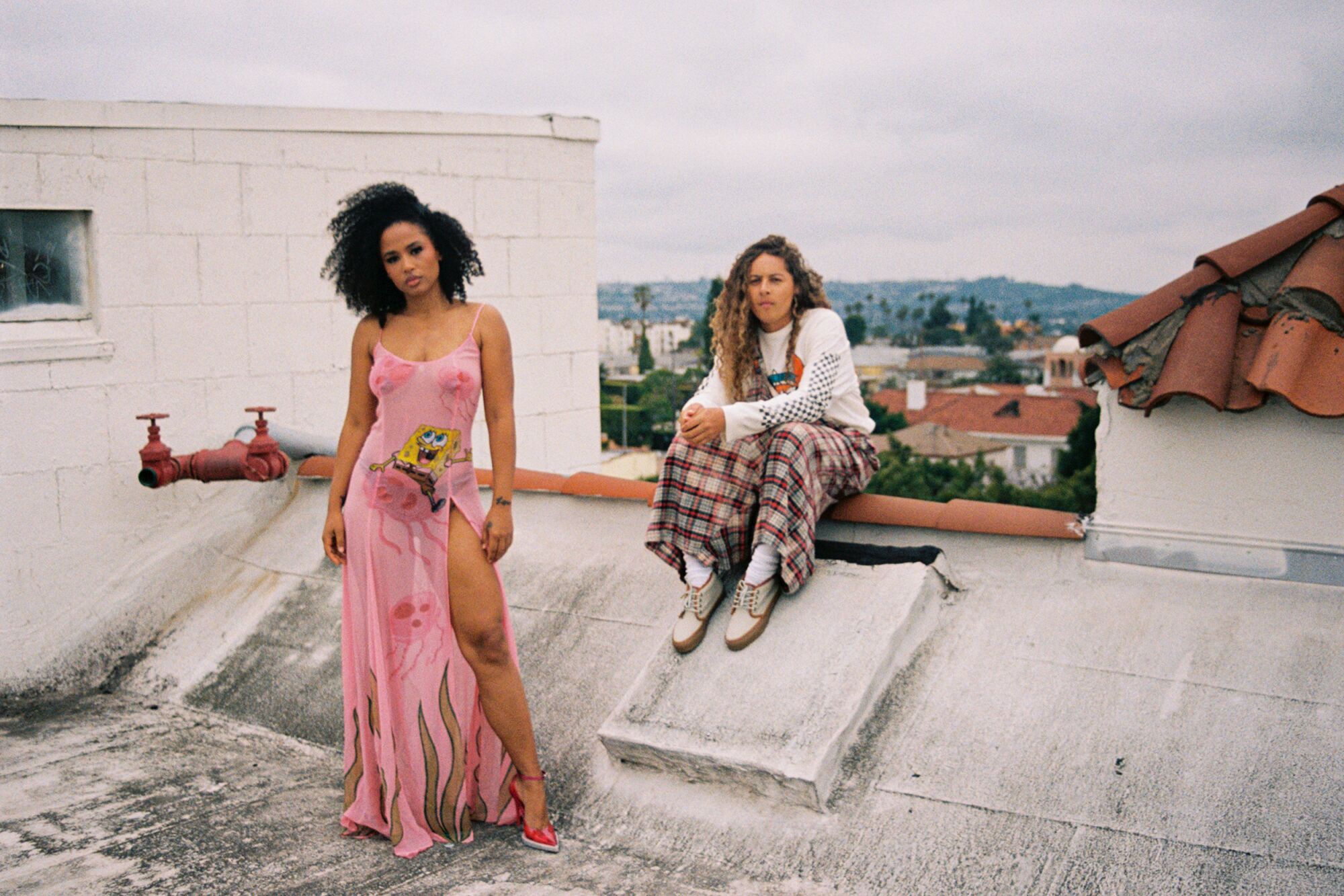 Two people on a residential rooftop, one standing wearing a pink dress and the other seated wearing plaid pants.
