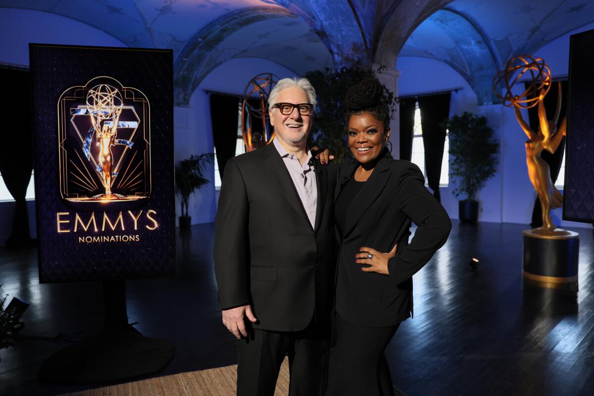 A man and a woman stand in a dramatically lit room with a sign that says Emmys behind them