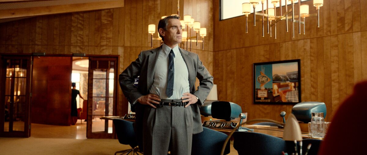 A man in a crisp suit stands, hands on hips, in a wood-paneled room.