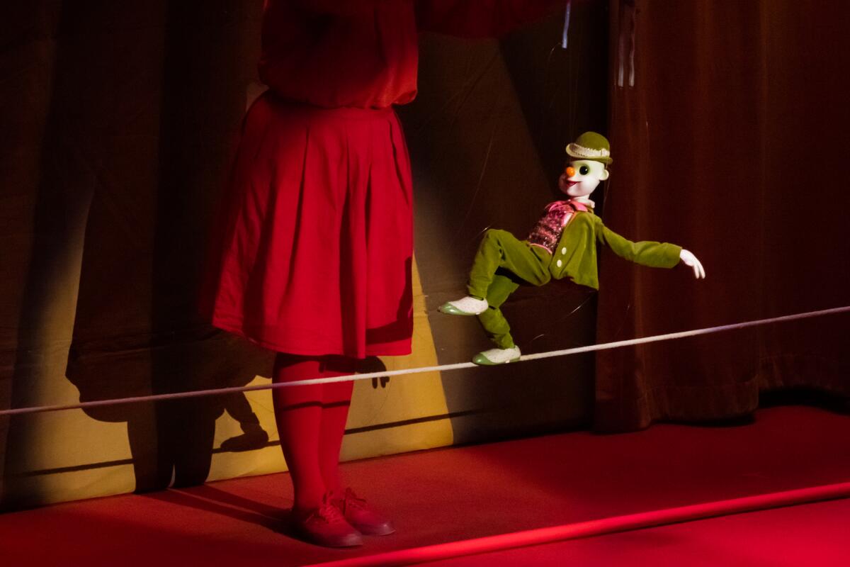 A marionette walks a high wire; just beyond, the puppeteer's red-skirted torso and legs are seen