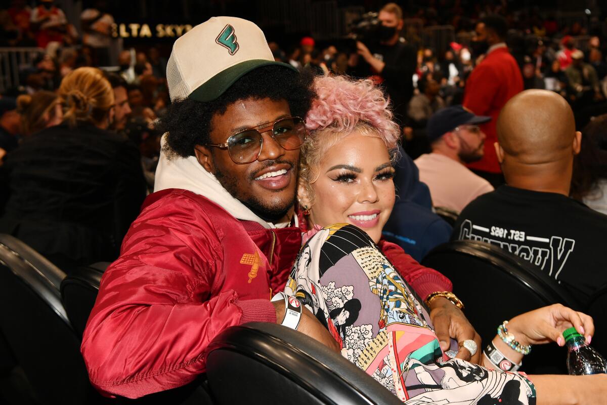 Jacky Oh, with pink hair, leans against her partner, DC Young Fly, who is wearing a red jacket, as they both sit smiling.