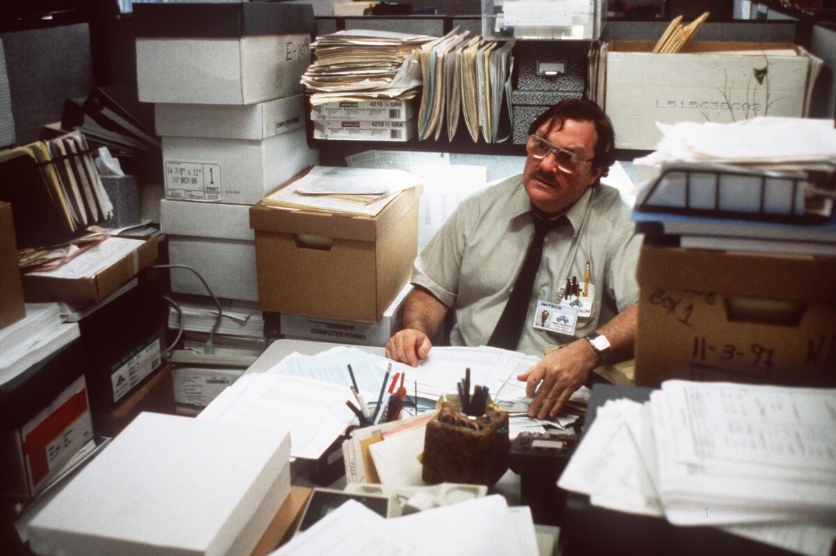 A man sits at a desk surrounded by banker boxes, stacks of paper and office supplies.